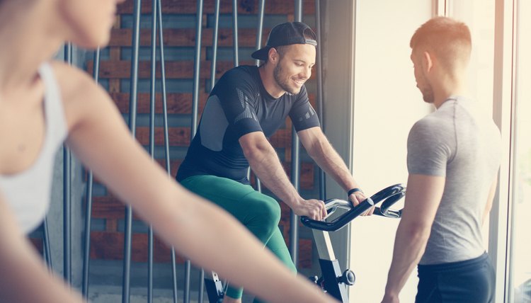 People exercise on stationary bikes in fitness class. People workout in gym. Exercise on elliptical machine. Man personal trainer.