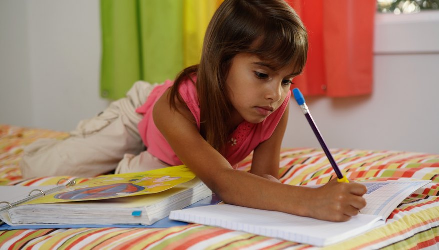 A young girl doing her homework while lying on a bed.