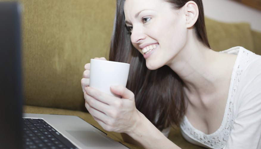 A woman holding a hot cup of tea while smiling at her computer screen.