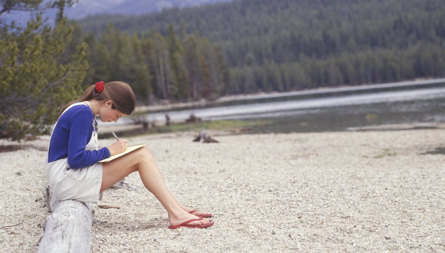 A teen sitting on a log and writing a letter by the lake in summertime.