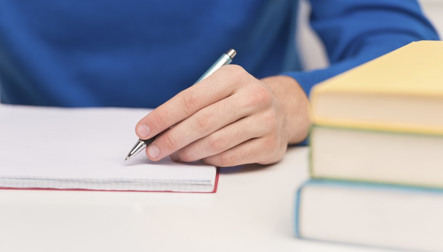 Student writing in notebook at table beside pile of books.