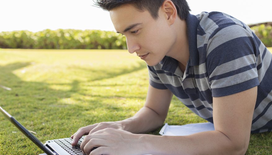 A college student typing on a laptop in an outdoor park.