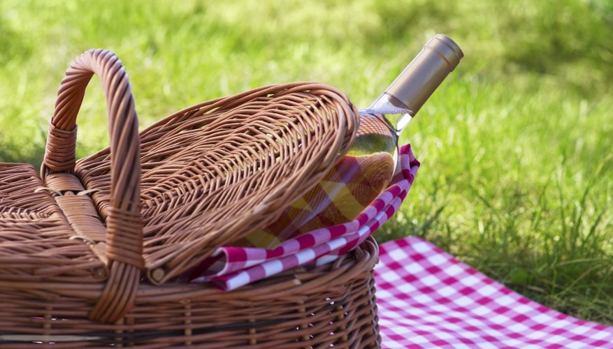 A bottle of white wine peeking out of a wicker basket on a red gingham tablecloth in the grass.