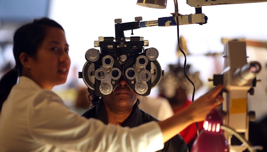 An optometrist performs an eye exam on a patient in a dim exam room.