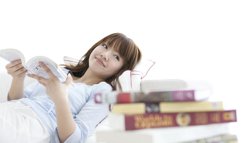 Young woman reading book in bed beside larger pile of books.