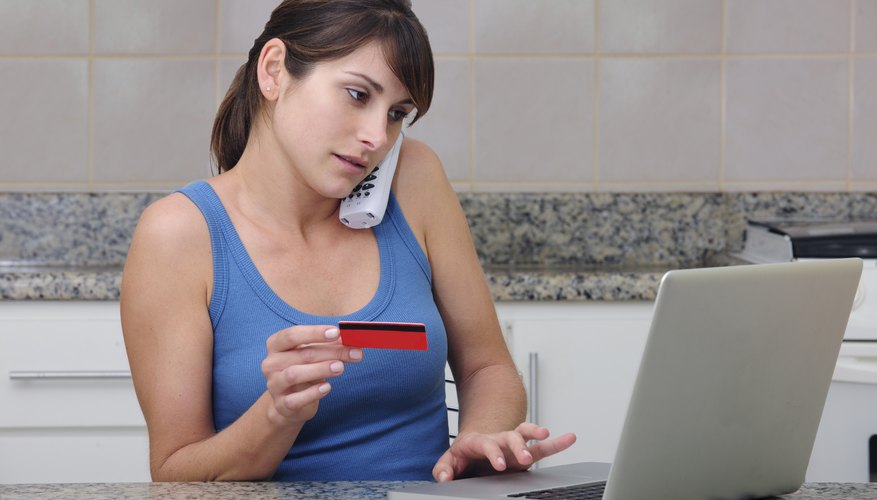 A young woman in her kitchen is on the phone with her credit card in hand and another hand on the keyboard of a laptop computer.