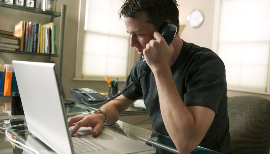 a young caucasian man sits working at his computer and on the phone in his home office