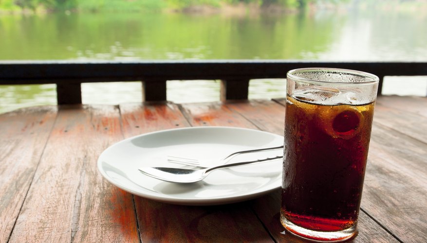 A glass of cola with ice on an outdoor table by a river. A glass of cola with ice on an outdoor table by a river.