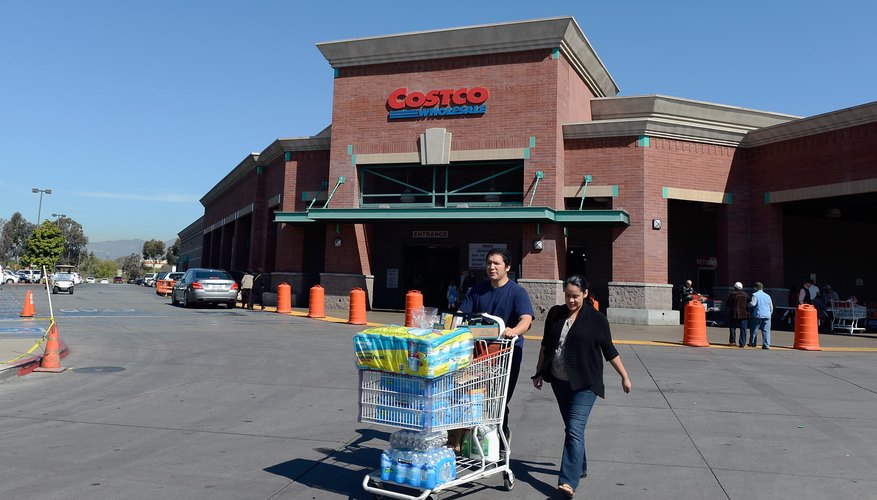 Consumers pushing a grocery cart of bulk items in the parking lot of a wholesale goods store.