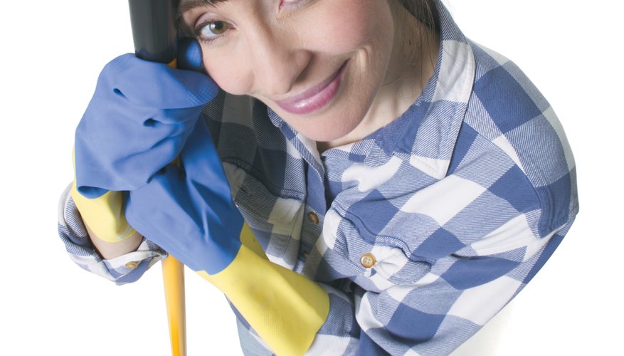 a caucasian woman in jeans and a plaid shirt wears rubber gloves and holds a mop as she looks up into the camera