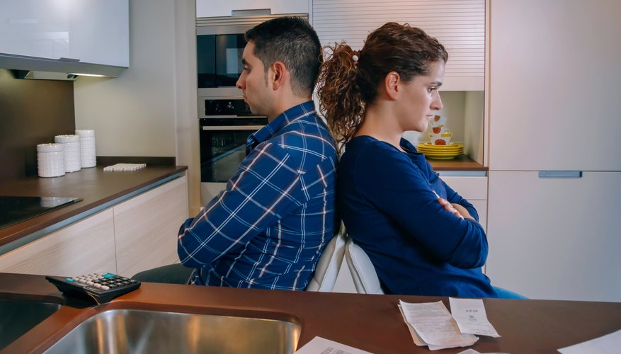 A couple sitting back to back in the kitchen with receipts on the counter.