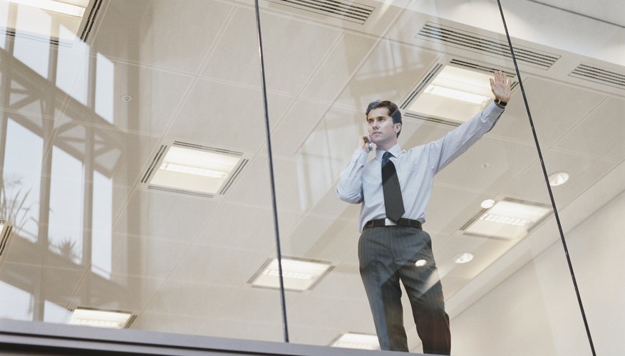 Businessman Looking Out of the Window of an Office Building and Using a Mobile Phone