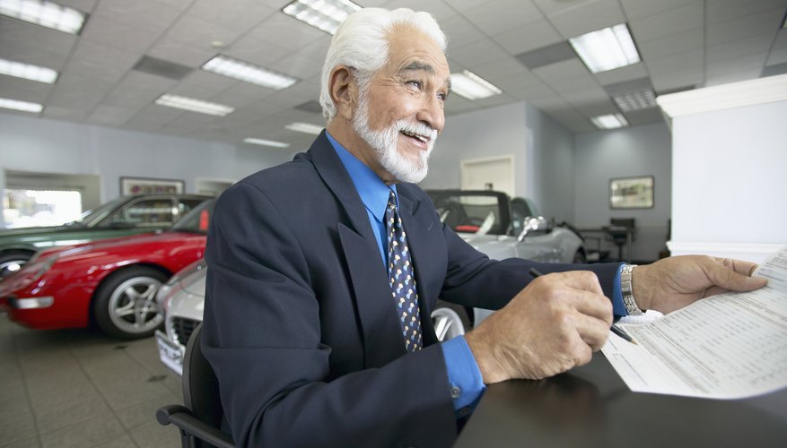 Senior Salesman Sits at a Table in a Car Showroom, Holding a Document