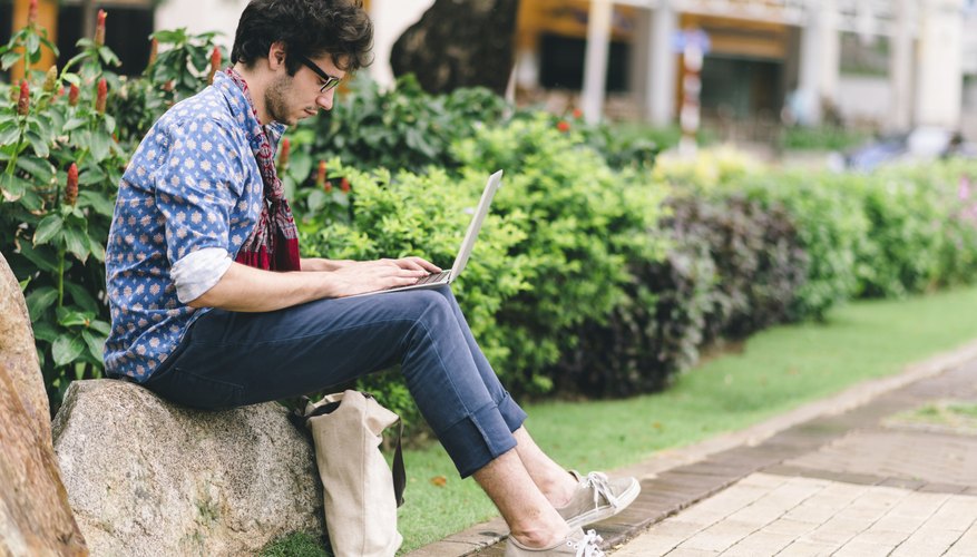 Photo Credit: DragonImages/iStock/Getty Images A young man perusing social media sites on his laptop in a courtyard.