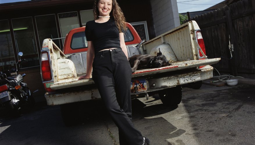 a young caucasian woman in black pants and shirt is leaning against the tailgate of a pickup truck with her black dog lying in the truck bed